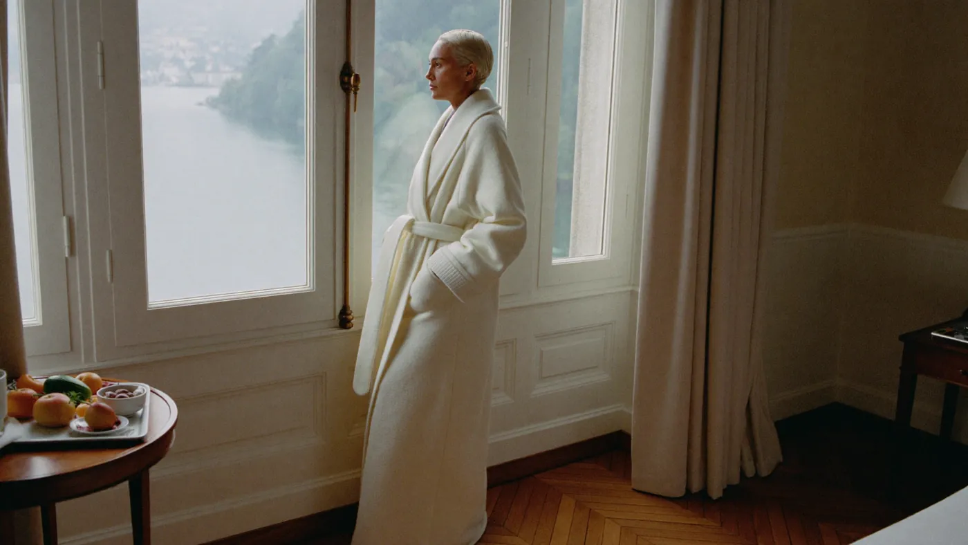 Eine Frau im Bademantel steht am hohen Fenster einer Hotelsuite am See, daneben ein Frühstückstablett.