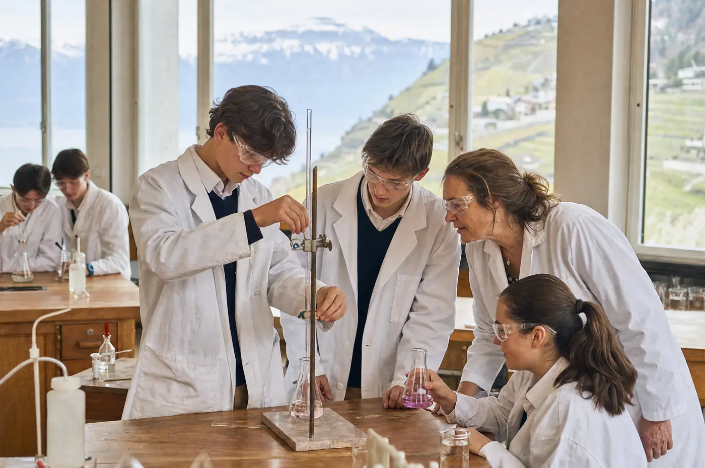Students in lab coats and goggles carry out an experiment with their chemistry teacher