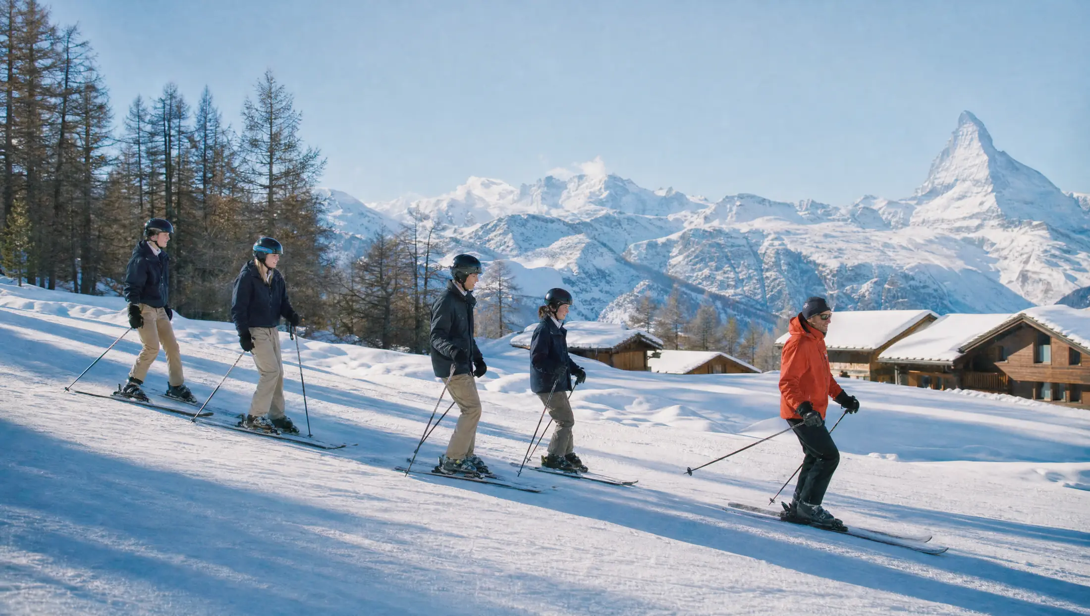 Students in helmets ski in a line behind their instructor on a sunny slope above wooden chalets